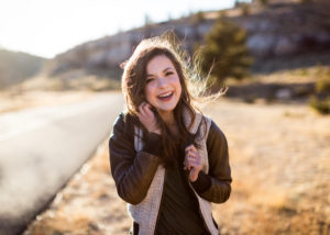 Senior girl wearing brown leather bomber jacket from Still In Style Boutique walking in field next to road laughing and holding her hair in the wind