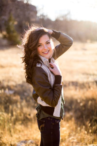 Senior girl wearing brown and white leather bomber jacket from Still In Style Boutique standing with left hand on collar and right hand running through her hair