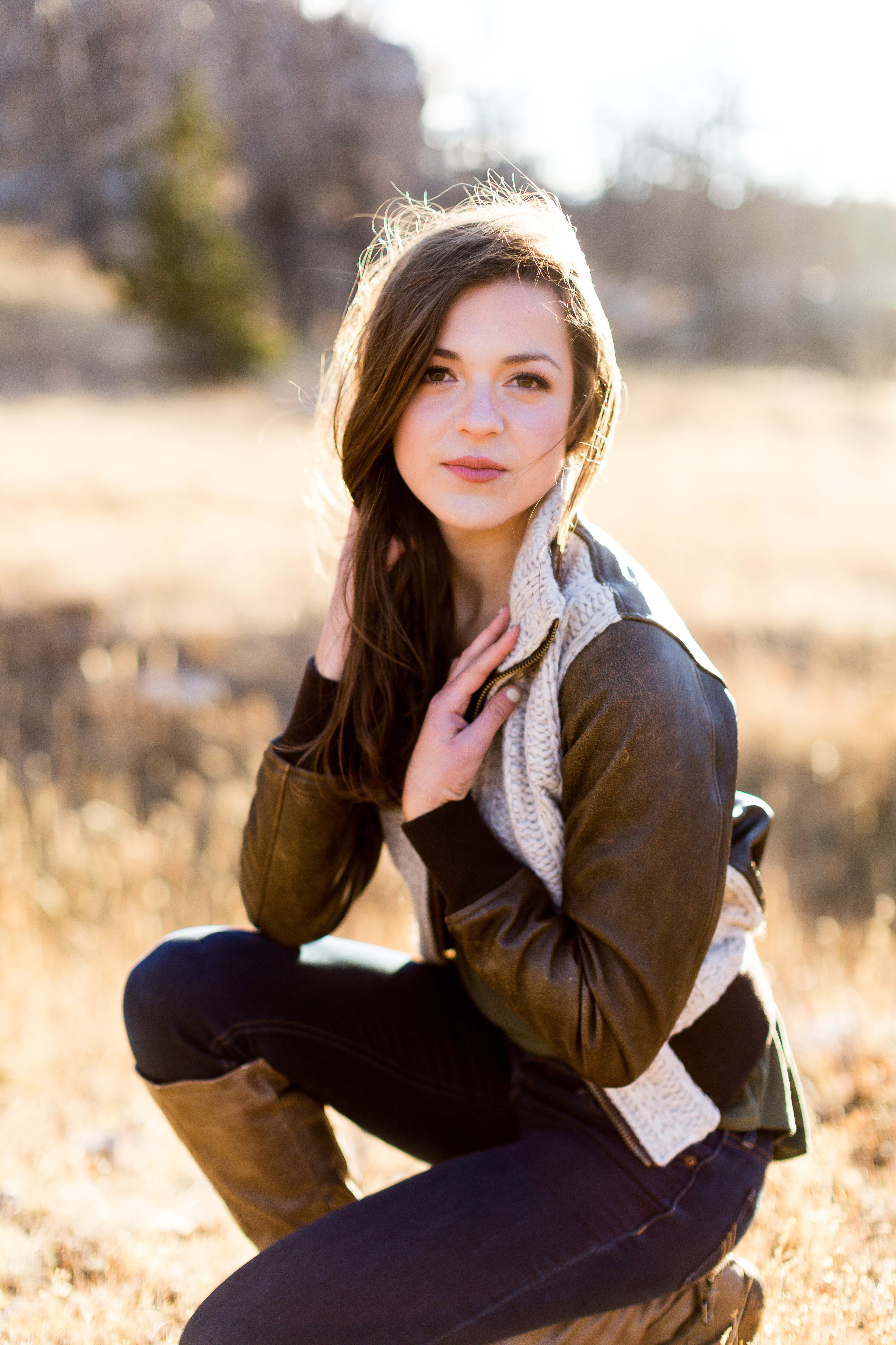 senior girl wearing brown and white leather bomber jacket from Still in Style Boutique kneeling in a field with her hands resting on collar