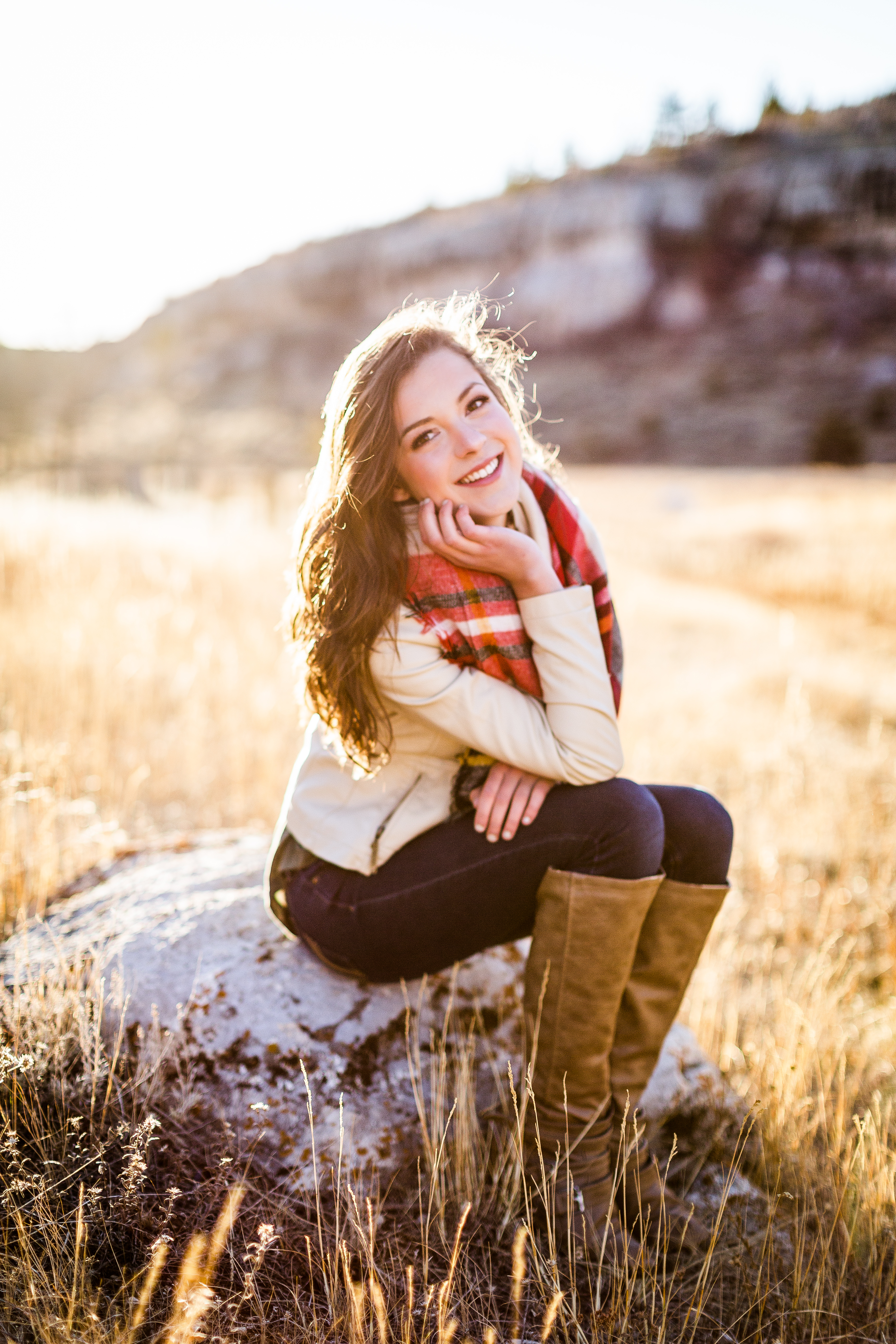 senior girl wearing a white leather jacket and red plaid balnket scarf sits on a rock in a field with her chin resting in her left hand and her right hand on her lap while smiling