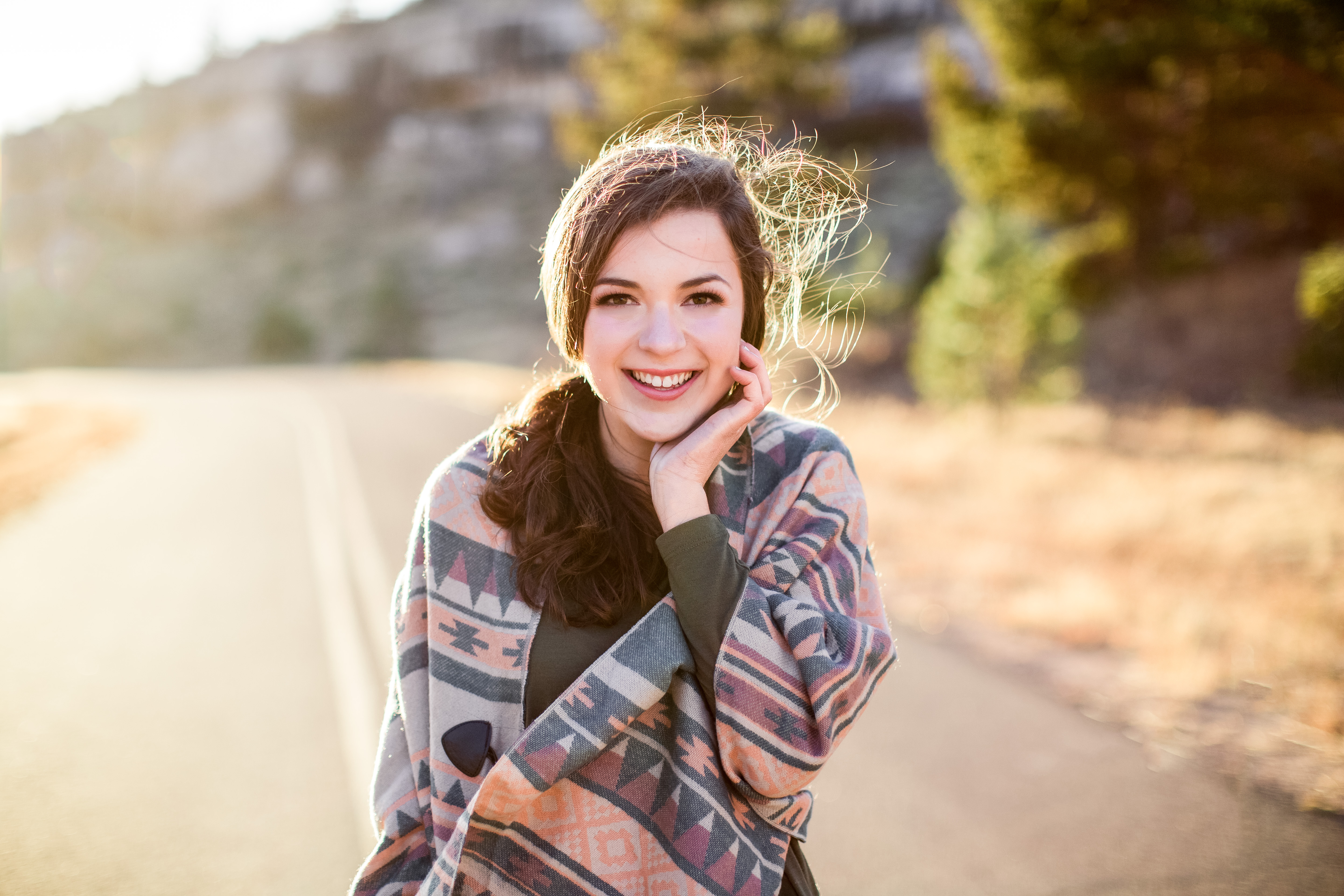 senior girl with brown hair wearing pink and teal patterned poncho from Still In Style Boutique stands smiling at the camera with her right hand caressing her cheek