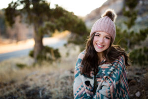 senior girl wearing pink hat with fur pom pom and pink and teal poncho from Still In Style Boutique sitting on the ground with her hands resting on her lap