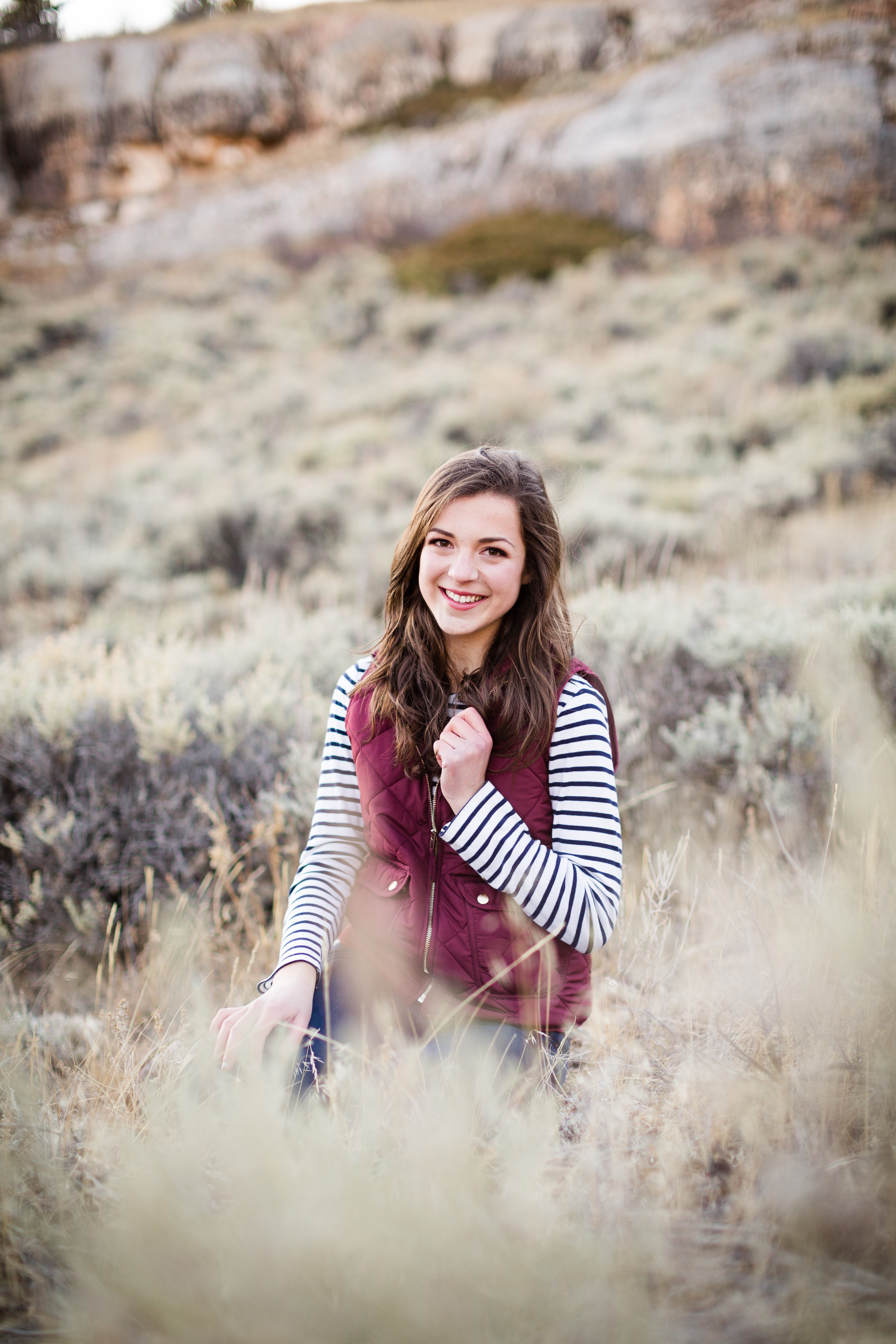 senior girl wearing a blue and white striped shirt and maroon vest sits in a field surrounded with sage brush with her right hand on her collar