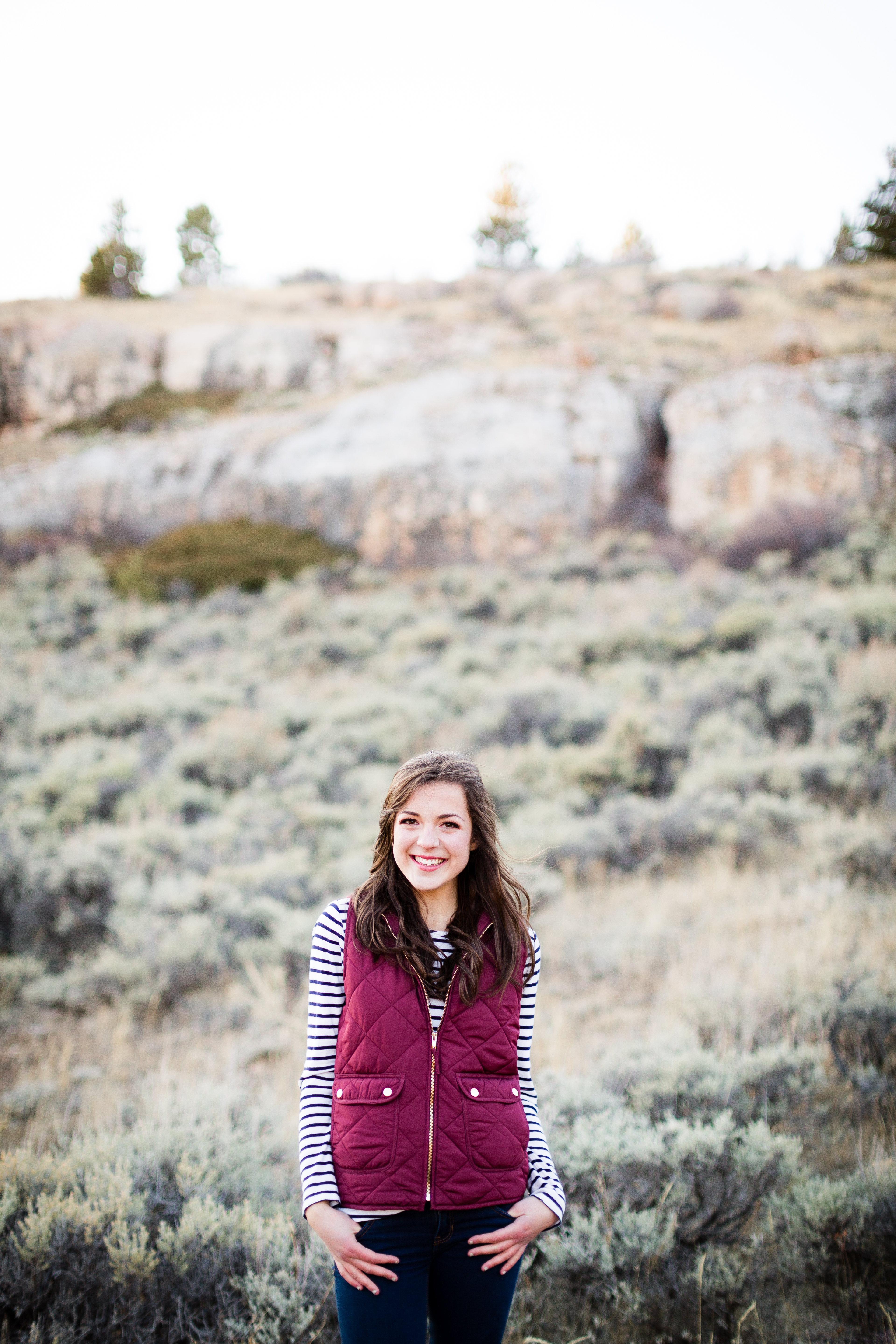 senior girl wearing a blue and white striped shirt and maroon vest stands smiling in a field surrounded by sage brush