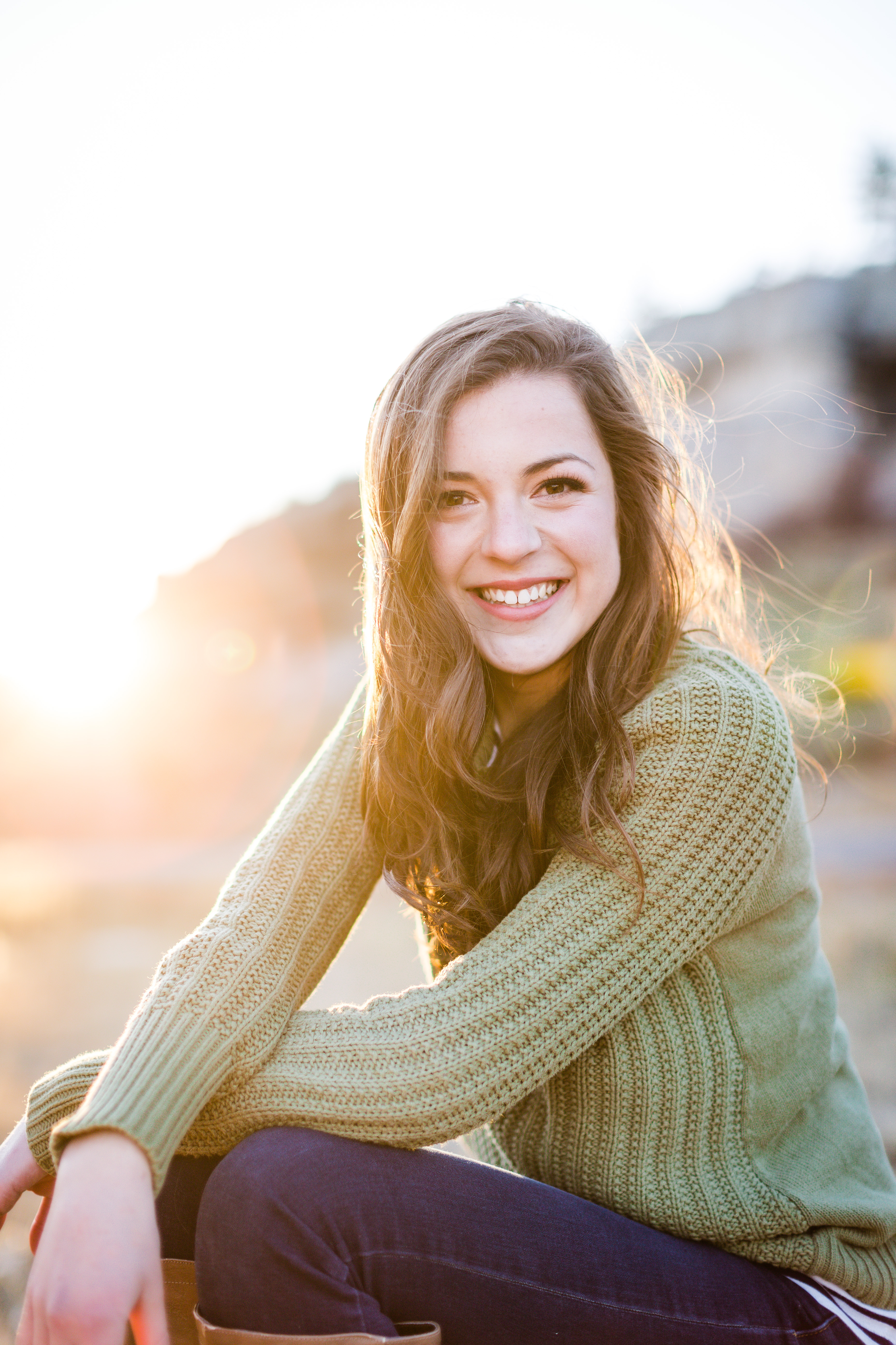 senior girl with long brown hair wearing a sage green sweater is smiling with her hands crossed over her knees