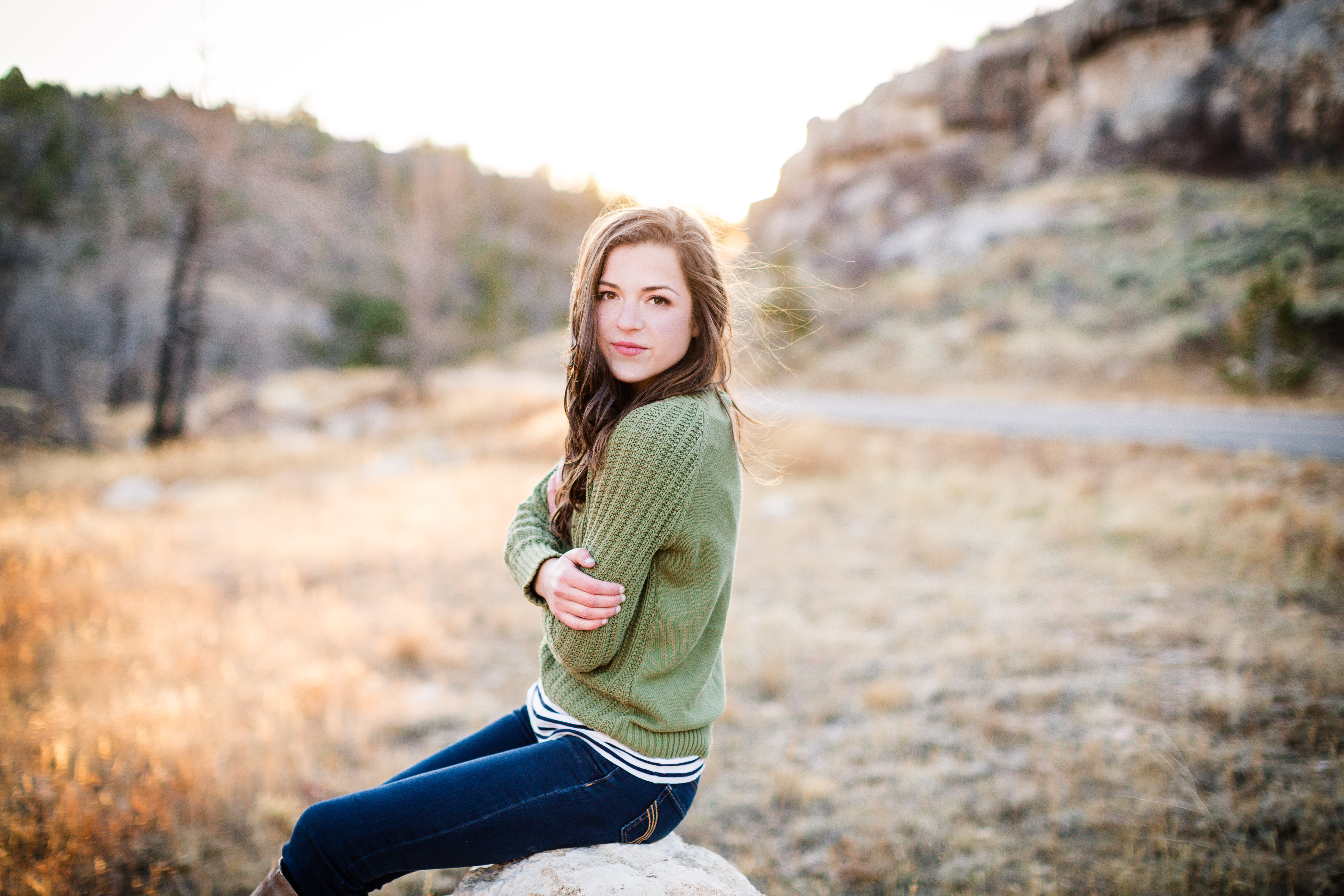 senior girl wearing sage green sweater and jeans sitting on a rock in a field with her arms crossed in front of her