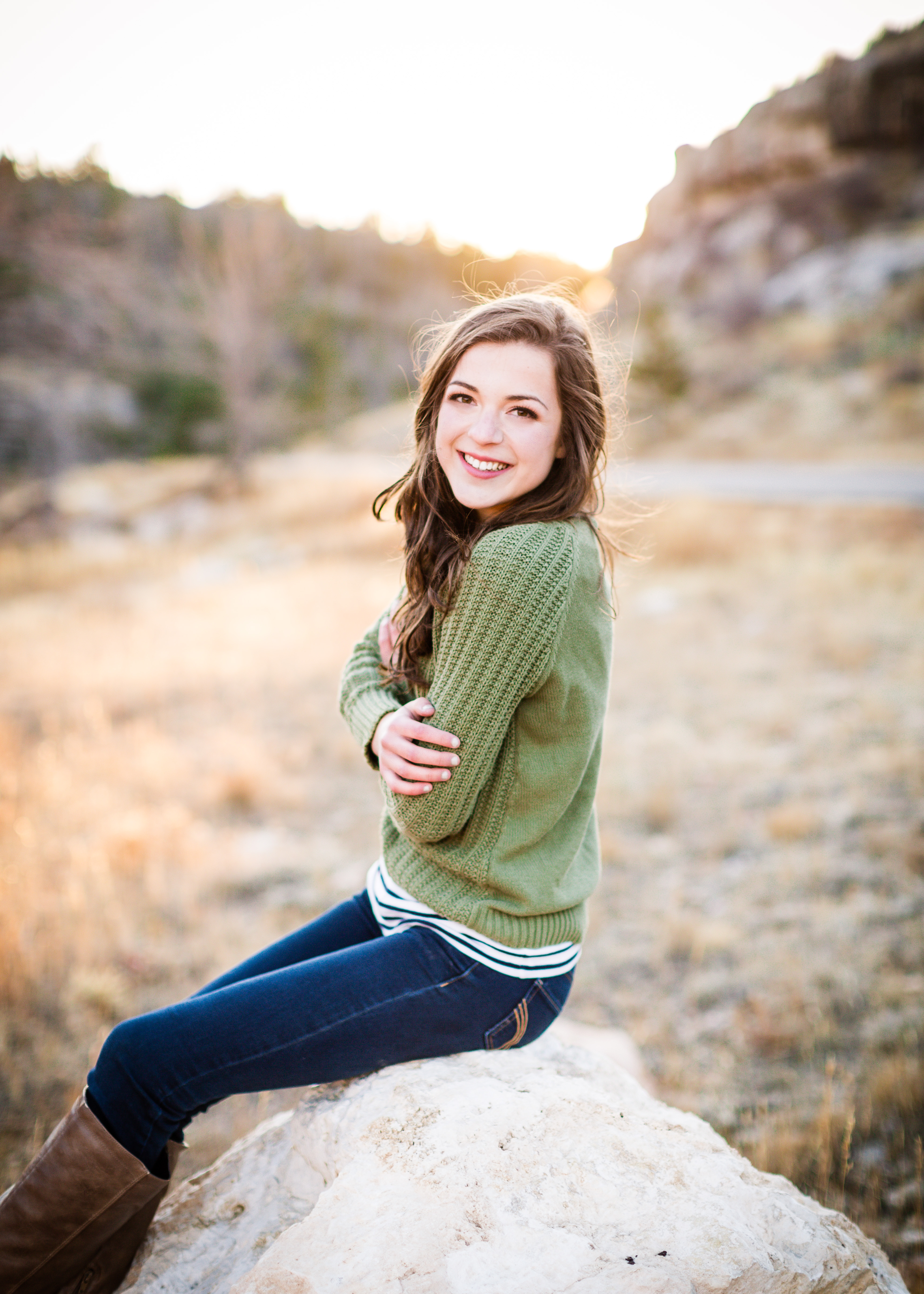 senior girl wearing sage green sweater and jeans sitting on a rock in a field smiling with her arms crossed in front of her