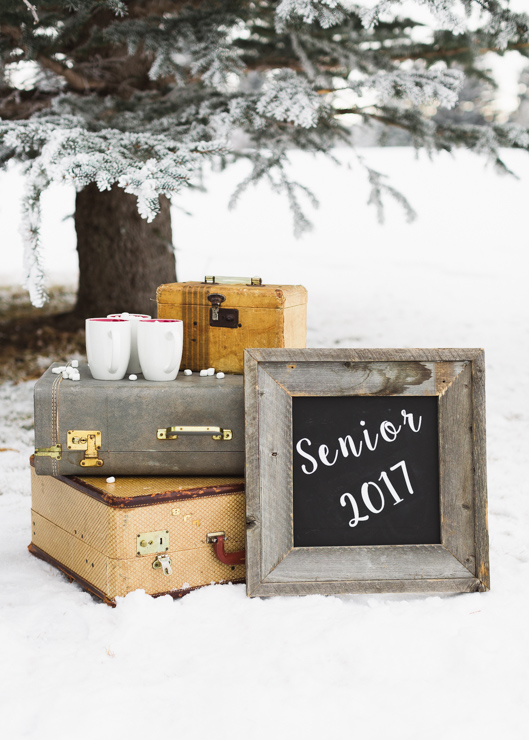 stacked vintage suitcases withe white mugs full of hot coco and chalkboard with senior 2017 written on it in the snow