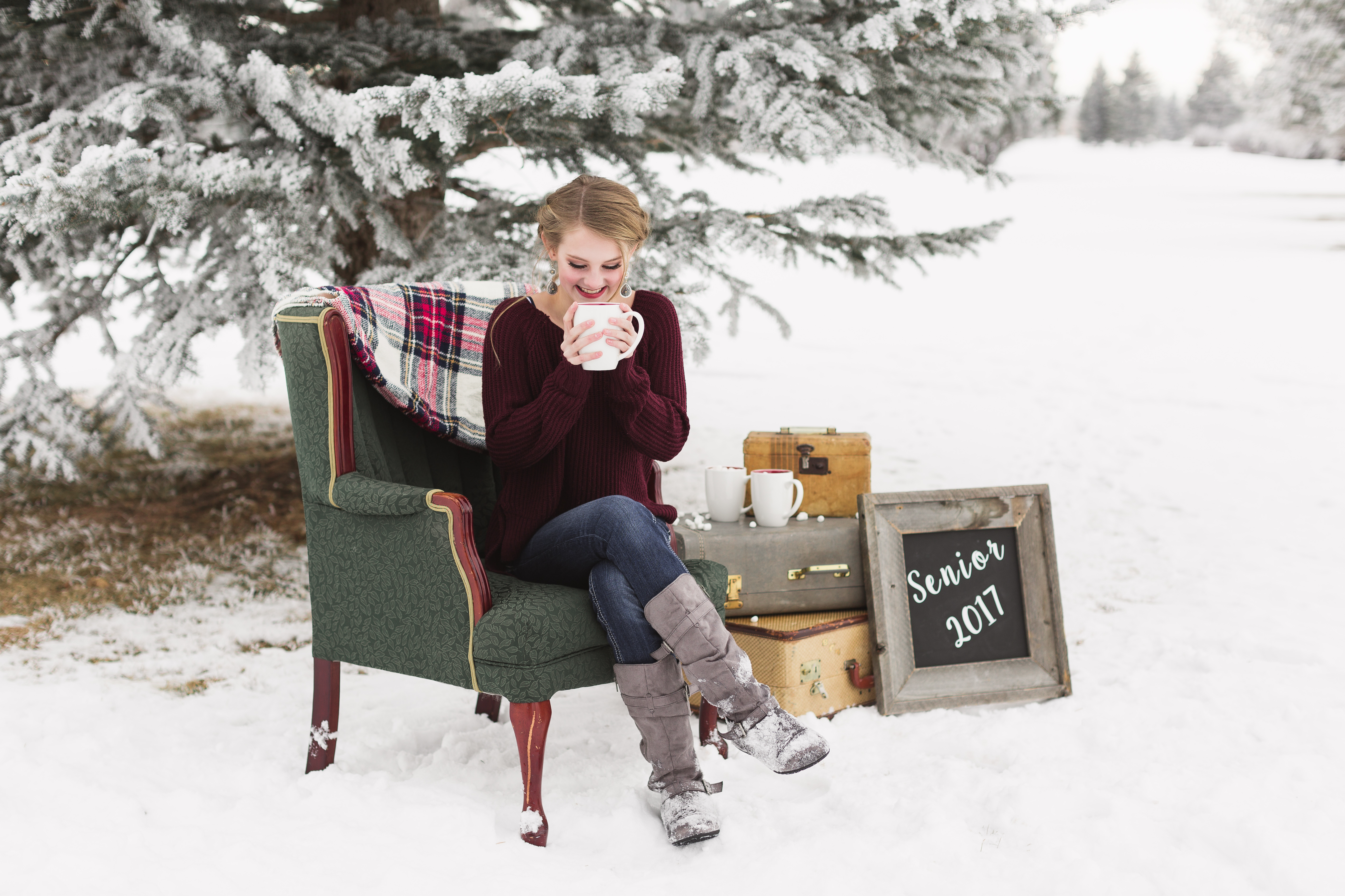 senior girl wearing maroon sweater sitting on green antique chair while sipping a white mug full of hot coco and marshmallows in the snow