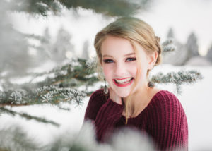 senior girl wearing maroon sweater smiling and is surrounded by branches of pine tree