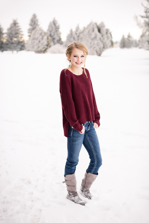 blonde senior girl wearig maroon sweater jeans and tall grey boots standing in snow smiling with hands in pockets