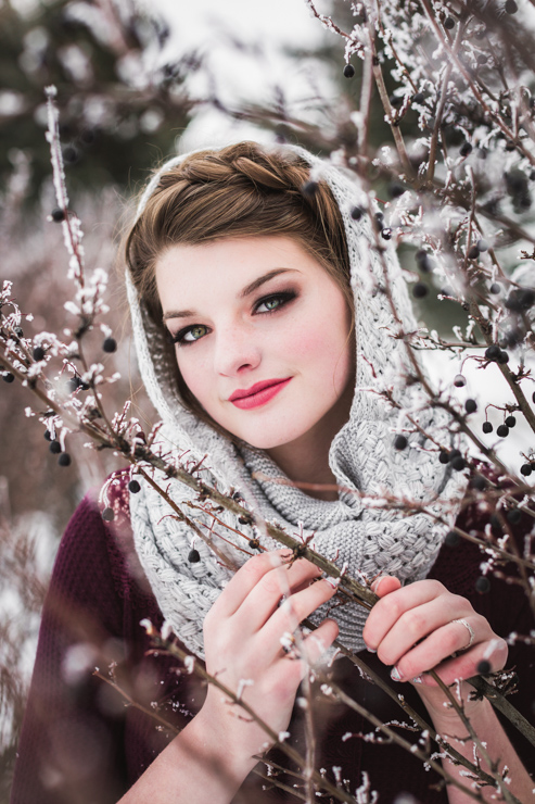 brunette senior girl with milkmaid braid and grey scarf draped on head is framed by branches of berry bush