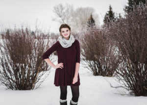senior girl wearing maroon dress, frey scarf, black tights and tall boots standing in snow with left hand on her hip