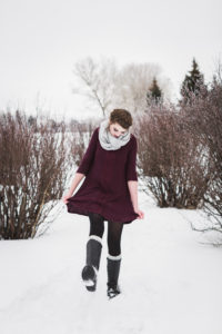 senior girl with brown hair in milkmaid braid wearing short maroon dress grey scarf and tall black boots kicking snow with left foot