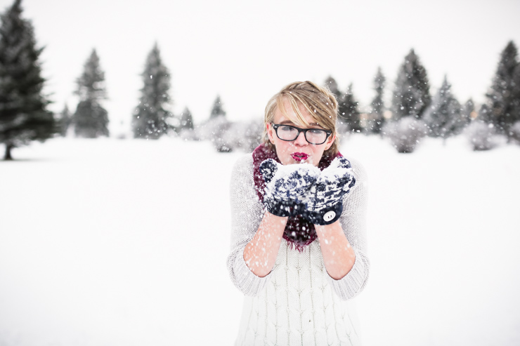 senior girl wearing white sweater maroon scarf and blue mittens blowing snow out of her hands