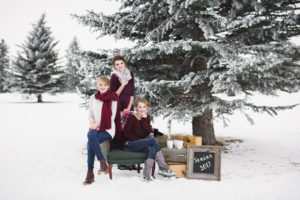 three senior girls posing around green antique chair , vintage suitcases, and chalkboard with senior 2017 written on it by pine tree in the snow