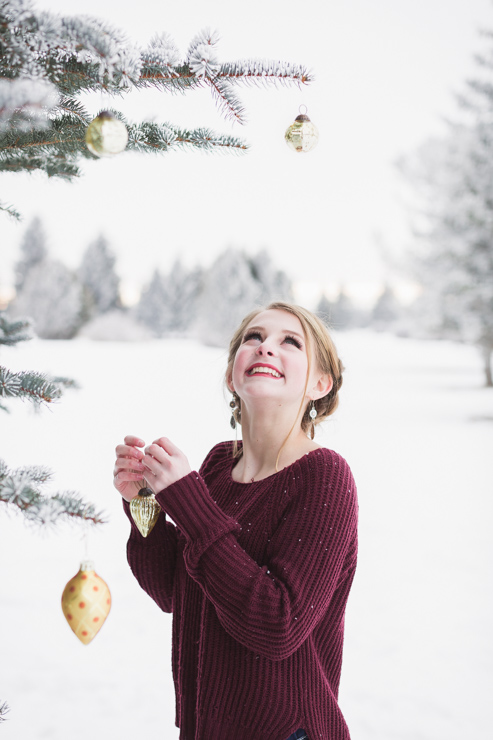 blonde senior girl in maroon sweater decorates pine tree with gold ornaments in the snow
