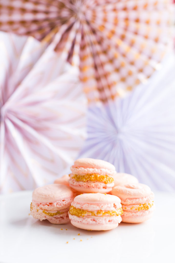 Pink and gold macaroons stacked on a white plate with pink and purple and gold paper fans in the background