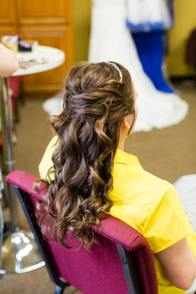 bride sits in a chair while a friend curls her long brown hair