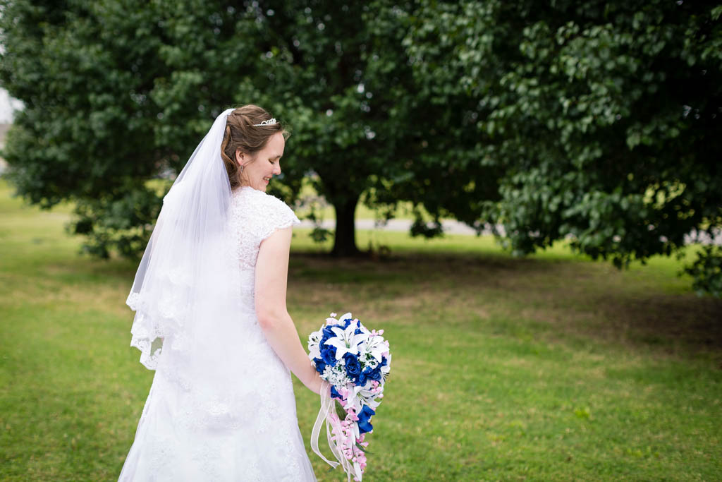 bride wearing a short sleeve lace dress and matching veil holds her blue, pink and white bouquet at her side and looks down at it