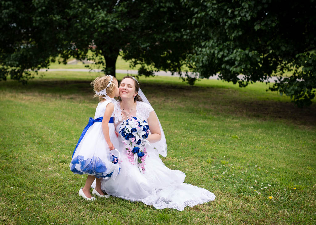 bride kneels down in the grass and smiles while her flower girl kisses her on the cheek