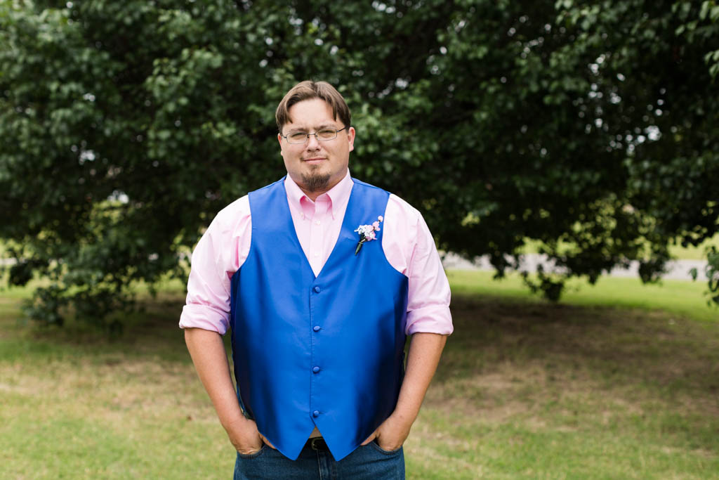 groom wearing pink shirt and royal blue vest smile with his hands tucked in his front pockets