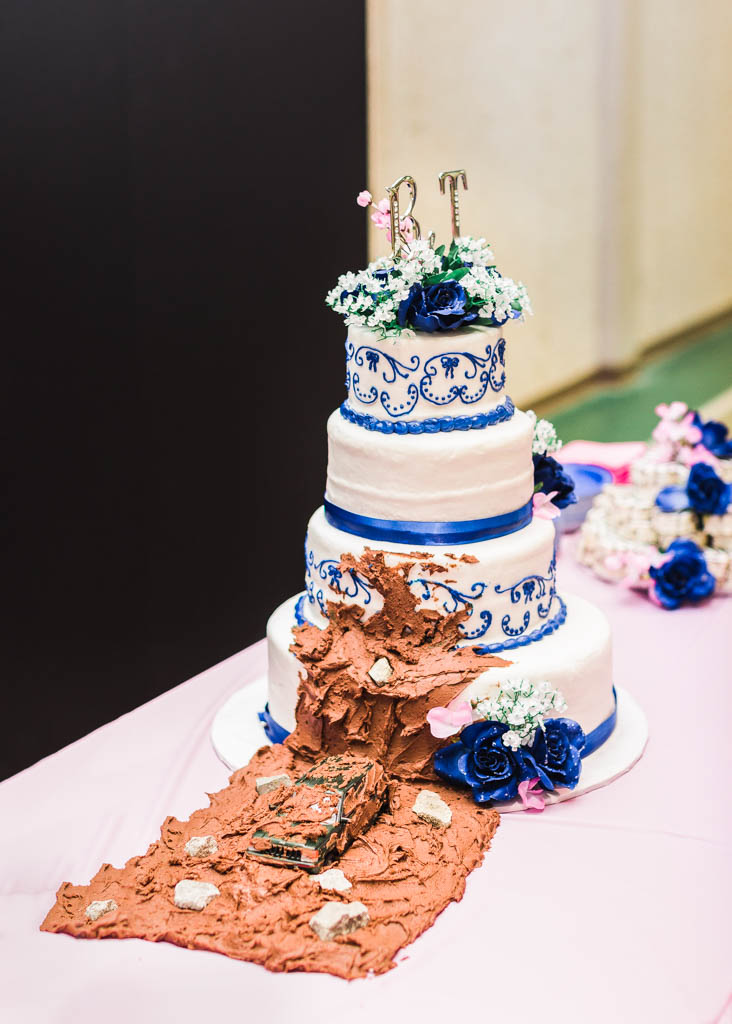 four tiered blue and white wedding cake with a patch of chocolate frosting and toy truck that looks like it is driving through mud on the side