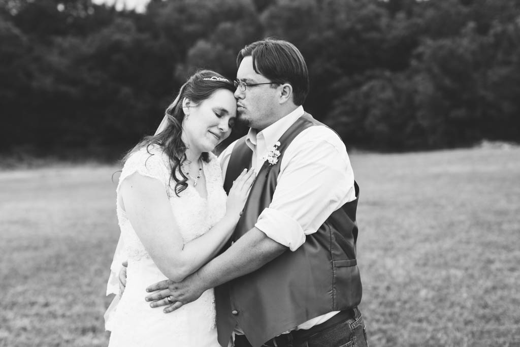 groom wraps his arms around his bride's waist and kisses her forehead in a field