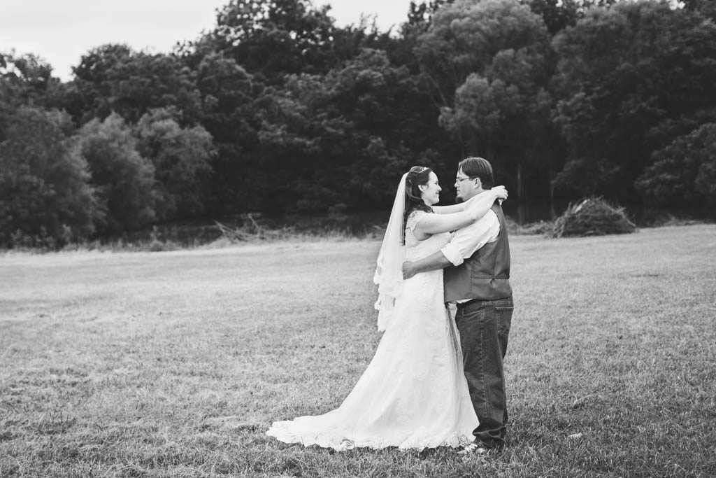 bride smiles and wraps her arms around her groom's neck in a field