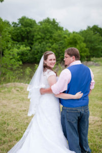 bride and groom wrap their arms around each other's backs and look over their shoulders at the camera