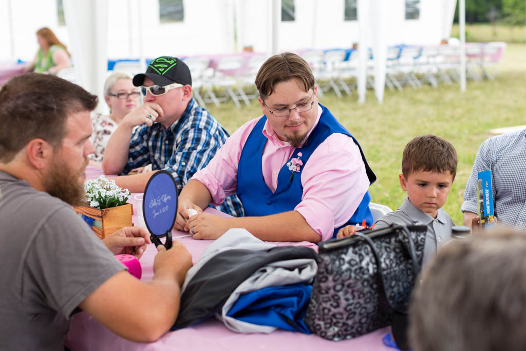 groom wearing a royal blue vest and pink shirt sits at a table and smiles at the ring bearer during the reception