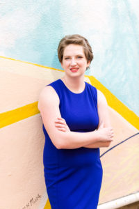 senior girl wearing a bright blue sleeveless dress leans against an abstract mural with her arms crossed