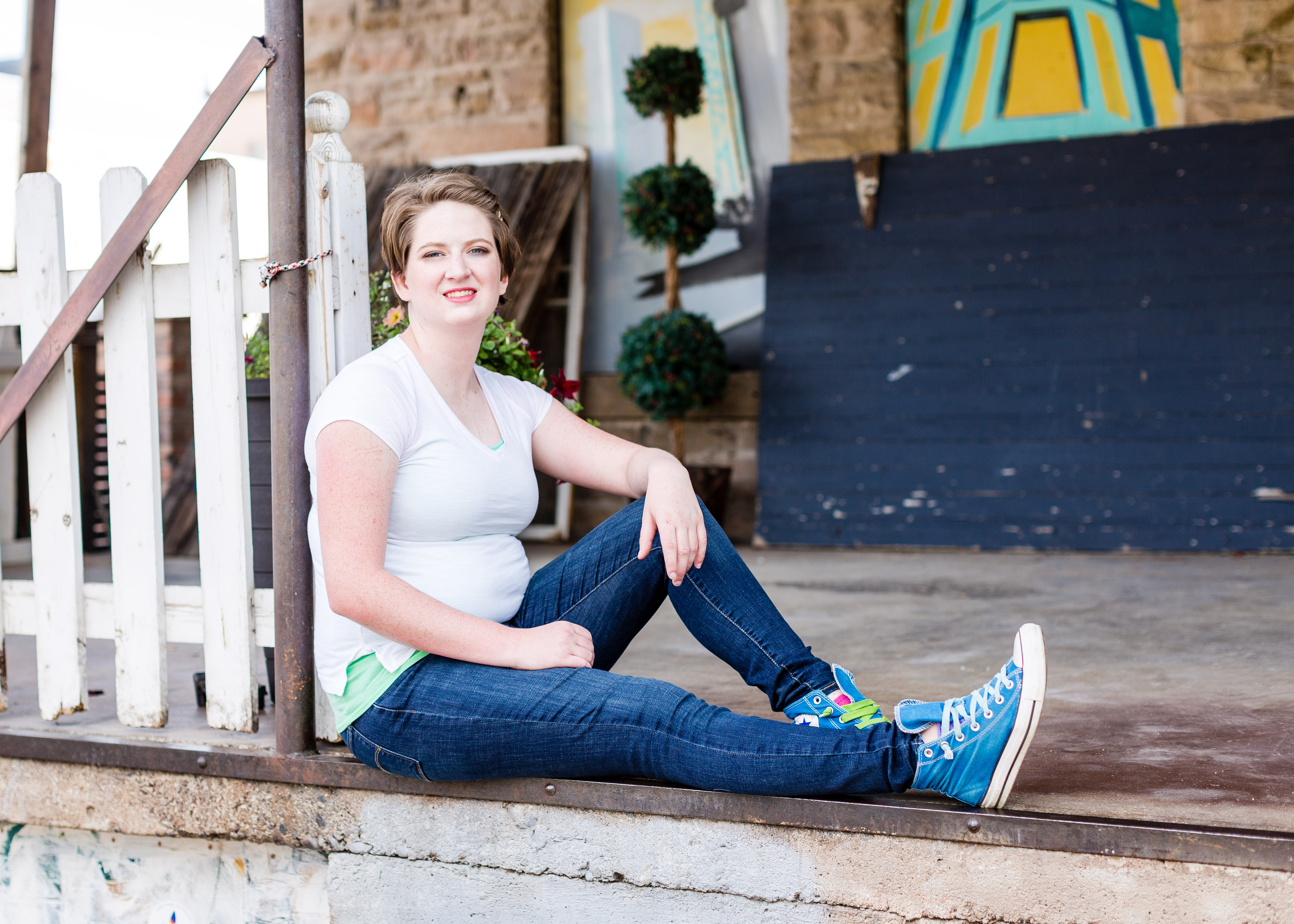 Senior girl wearing a white t-shirt and blue jeans sits with one leg bent and the other extended in a colorful alley