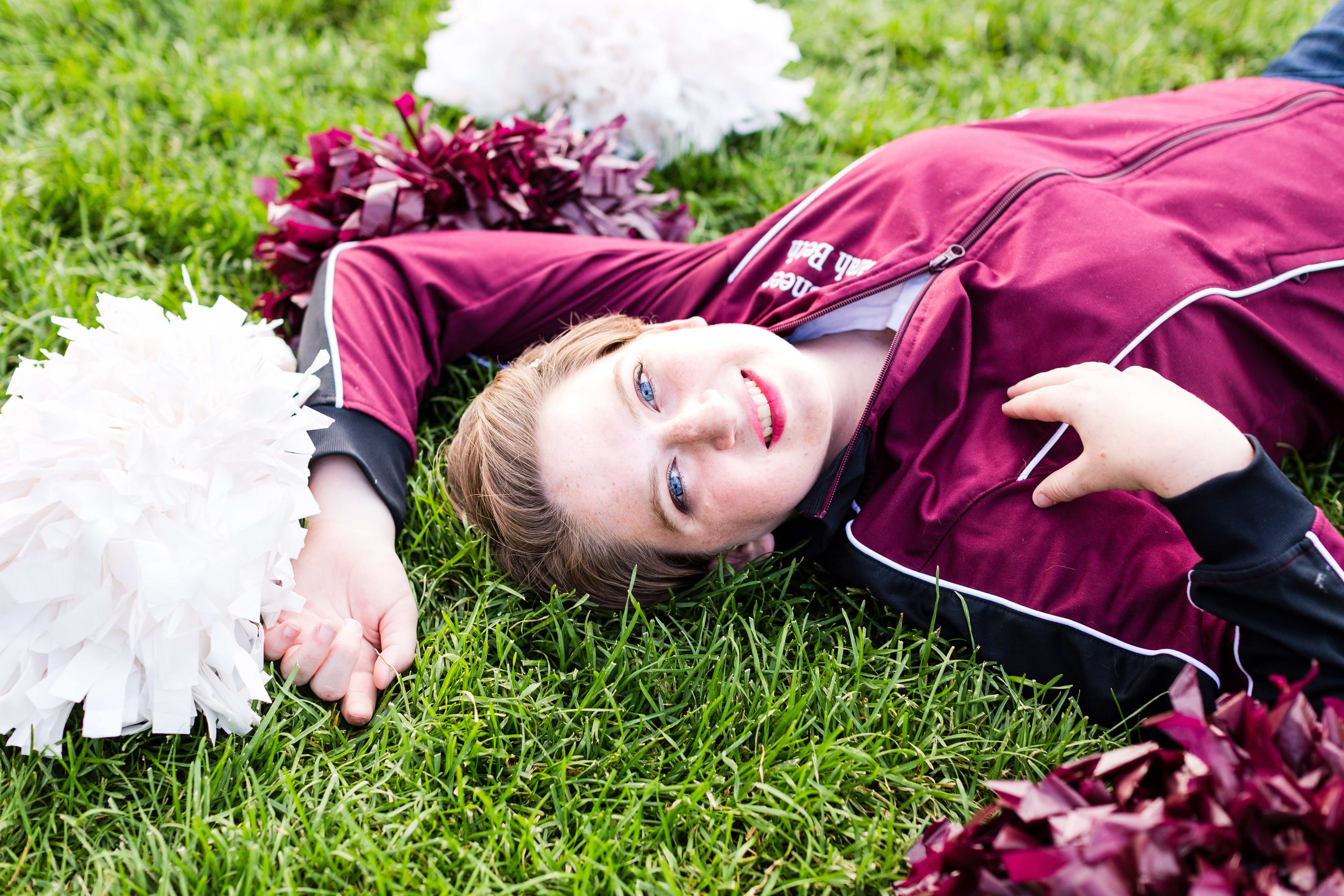senior girl wearing a maroon and black cheerleader jacket lays in the grass surrounded by her maroon and white pom poms