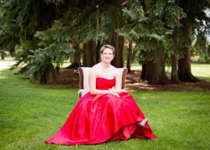 senior girl wearing a bright red strapless prom dress sits in an antique white armchair with pine trees in the background