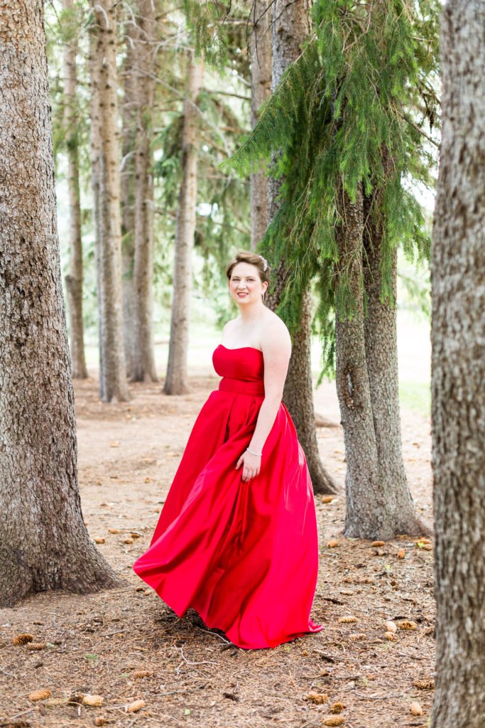 Senior girl short hair wearing a bright red strapless prom dress stands amongst pine trees