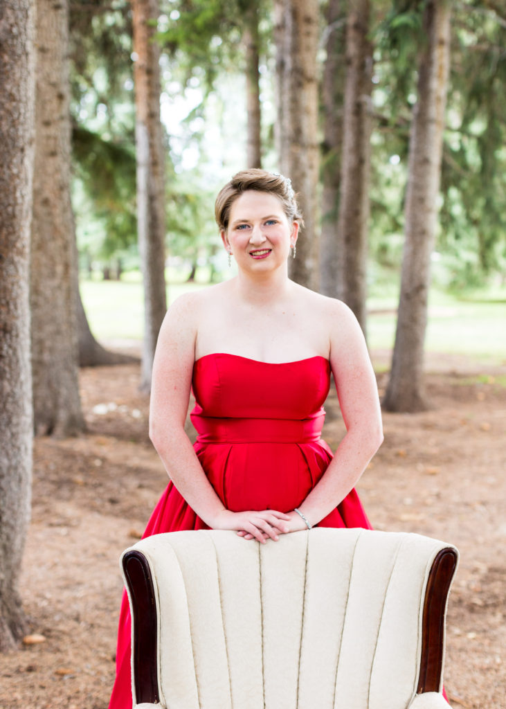 senior girl wearing bright red dress stands behind a white antique armchair amongst pine trees