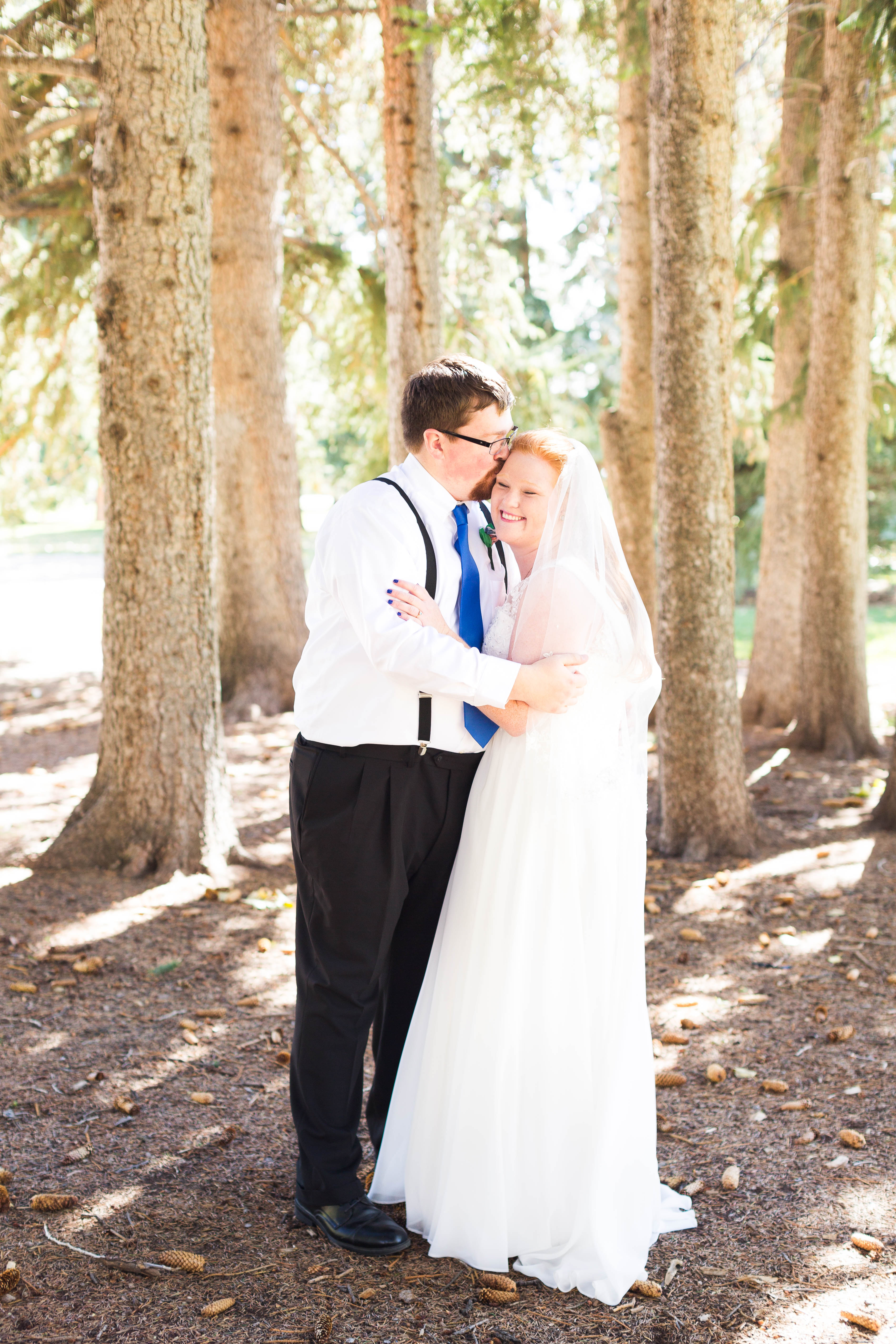 groom kisses his bride's forehead they embrace amongst pine trees
