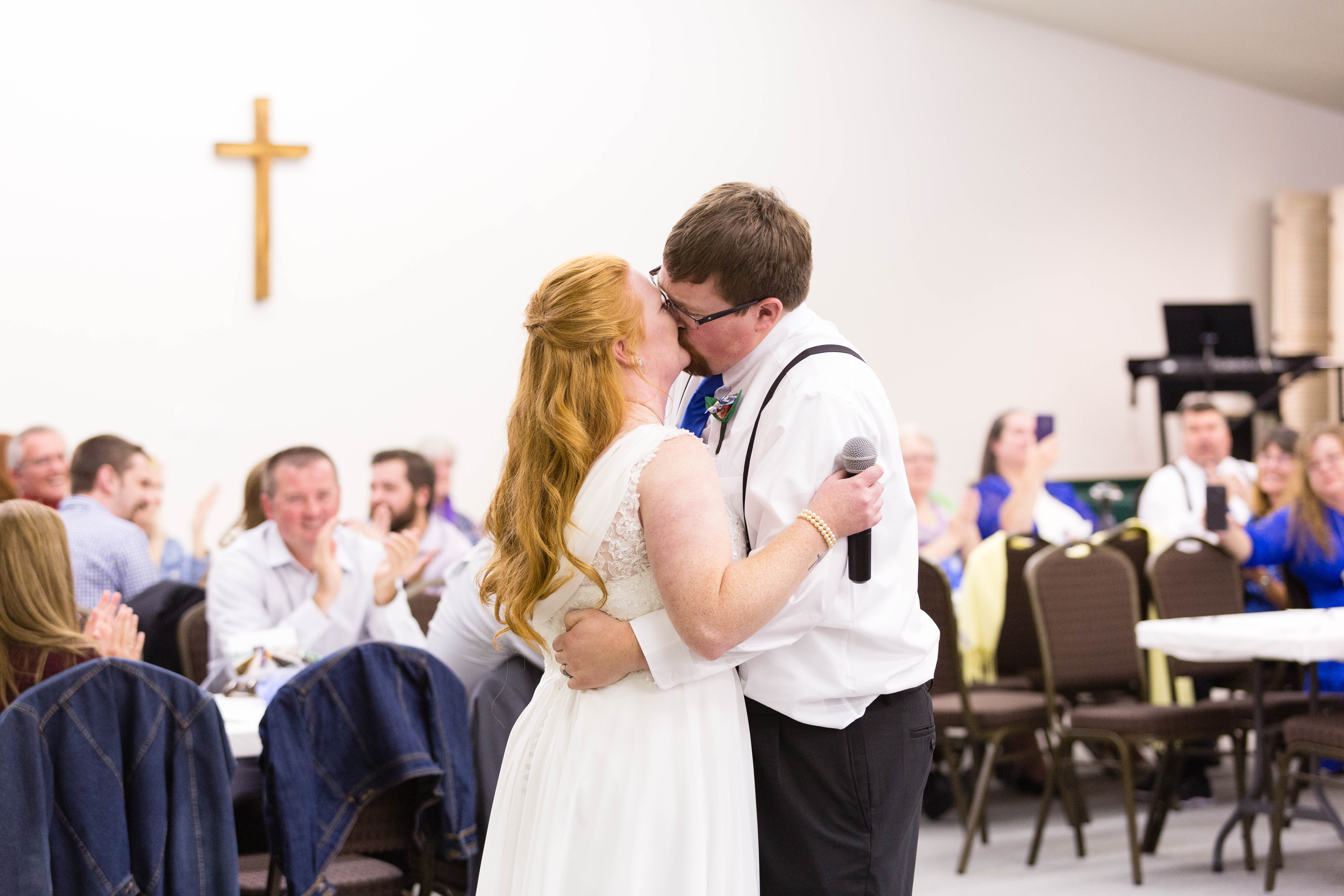 bride and groom kiss while guests applaud in the background at their reception