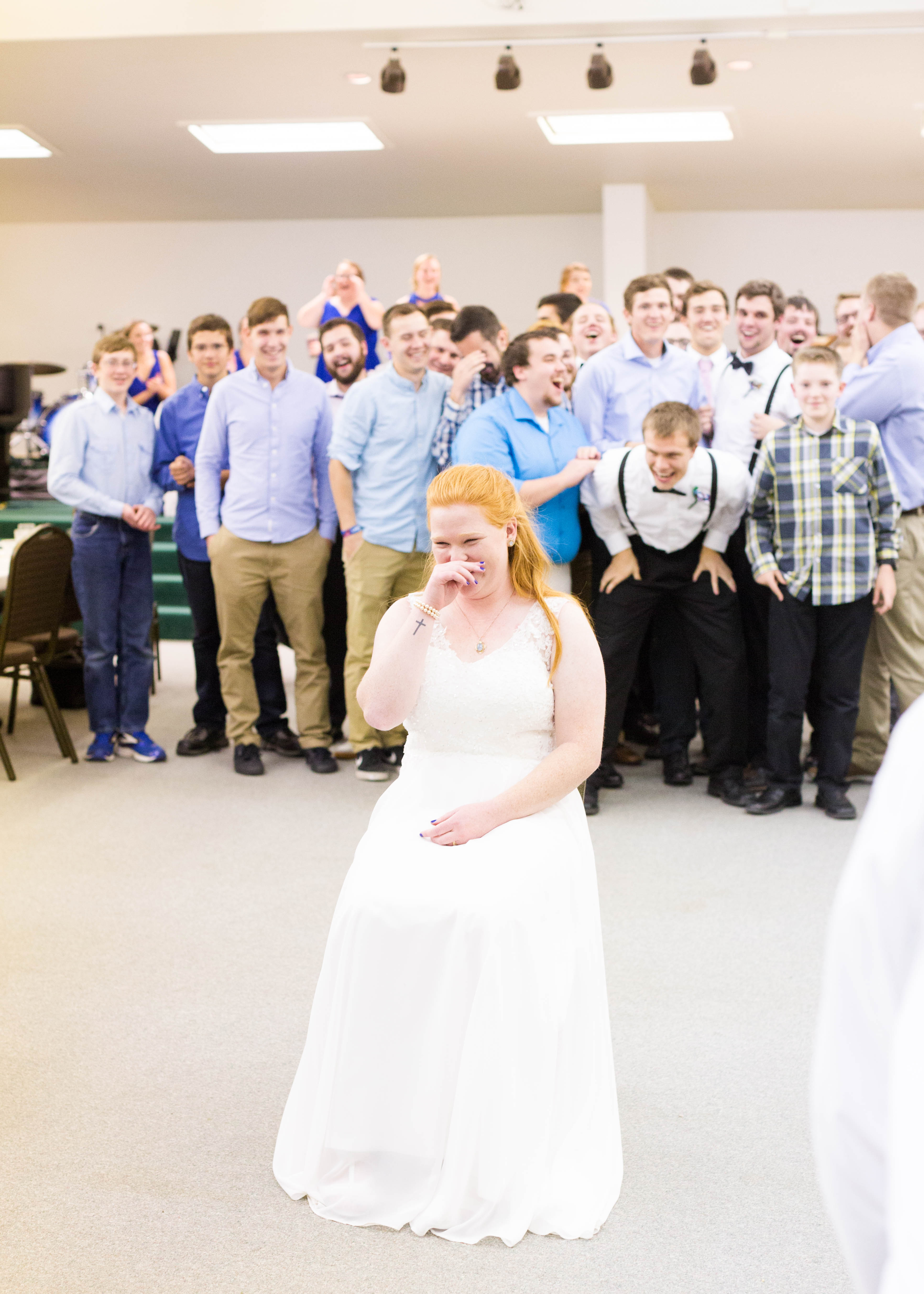 bride blushes and laughs while single men gather behind her for the garter toss