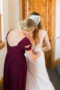 Bridesmaid in burgundy dress helps bride button her dress