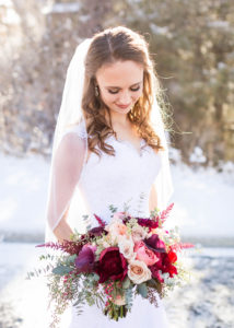 bride stands in the snow and gazes down at her pink and burgundy bouquet