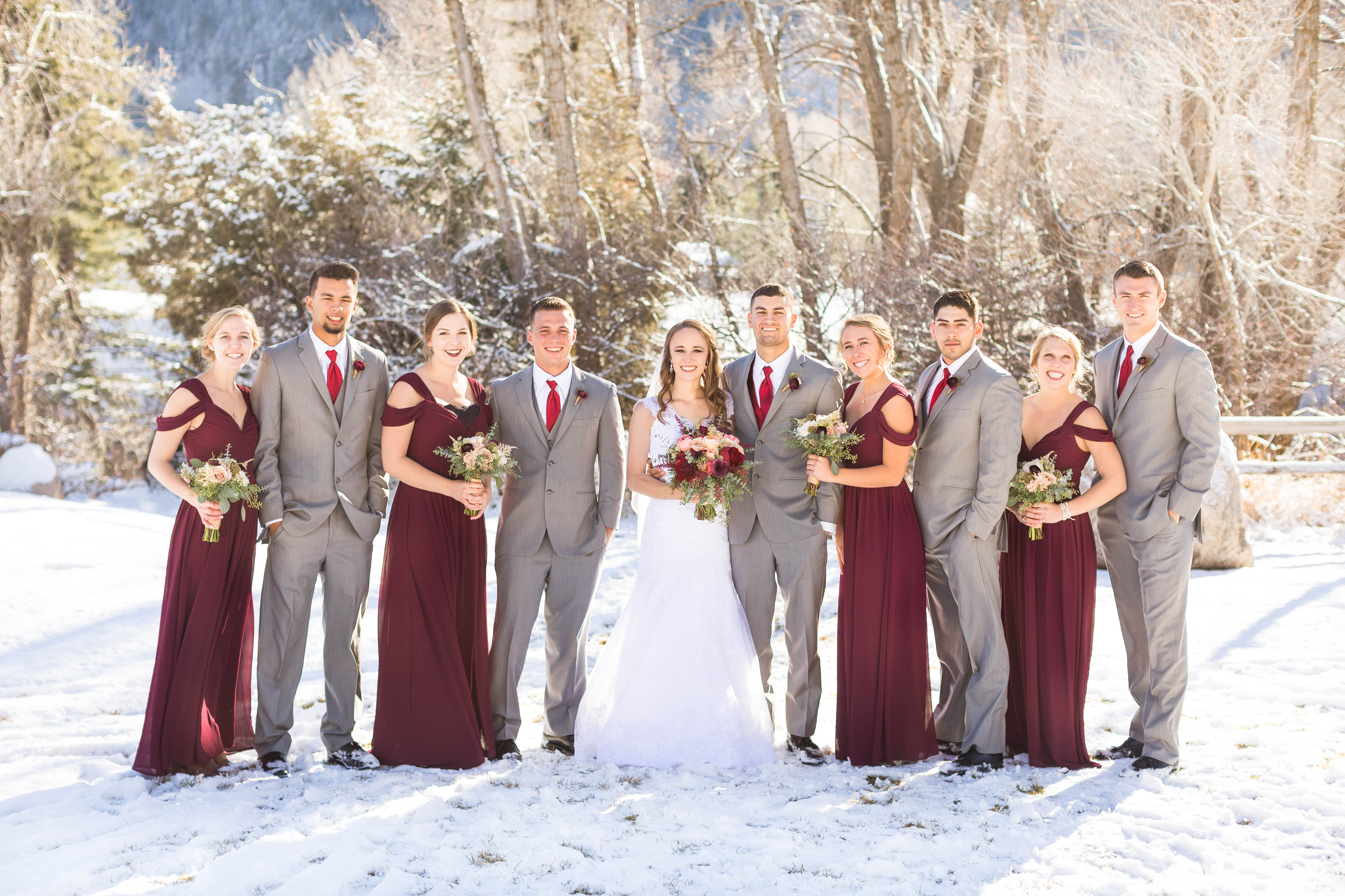 wedding party in burgundy dresses and grey suits gather around couple
