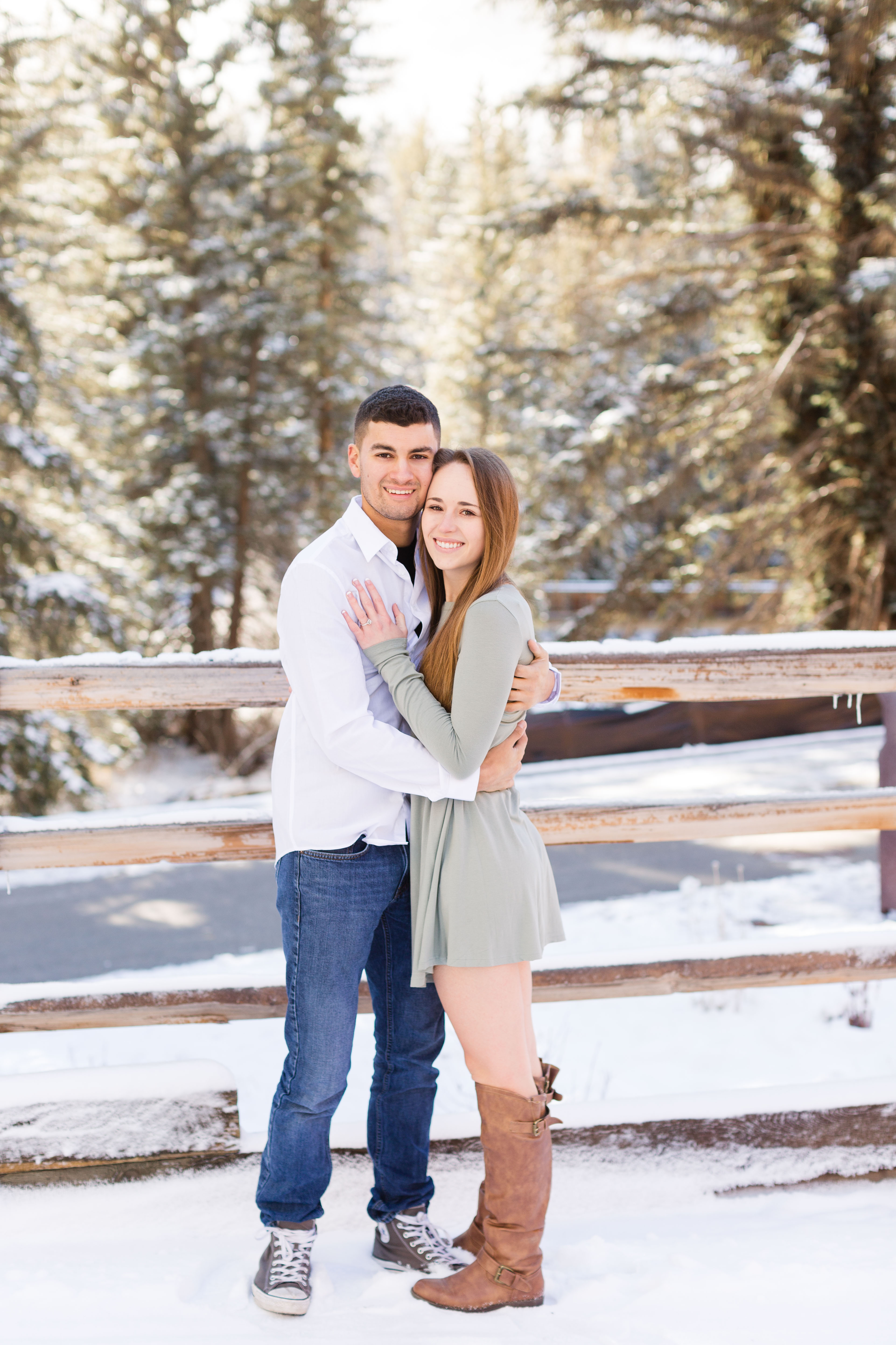 A couple hold each other and smile in front of a snow covered fence and pine trees