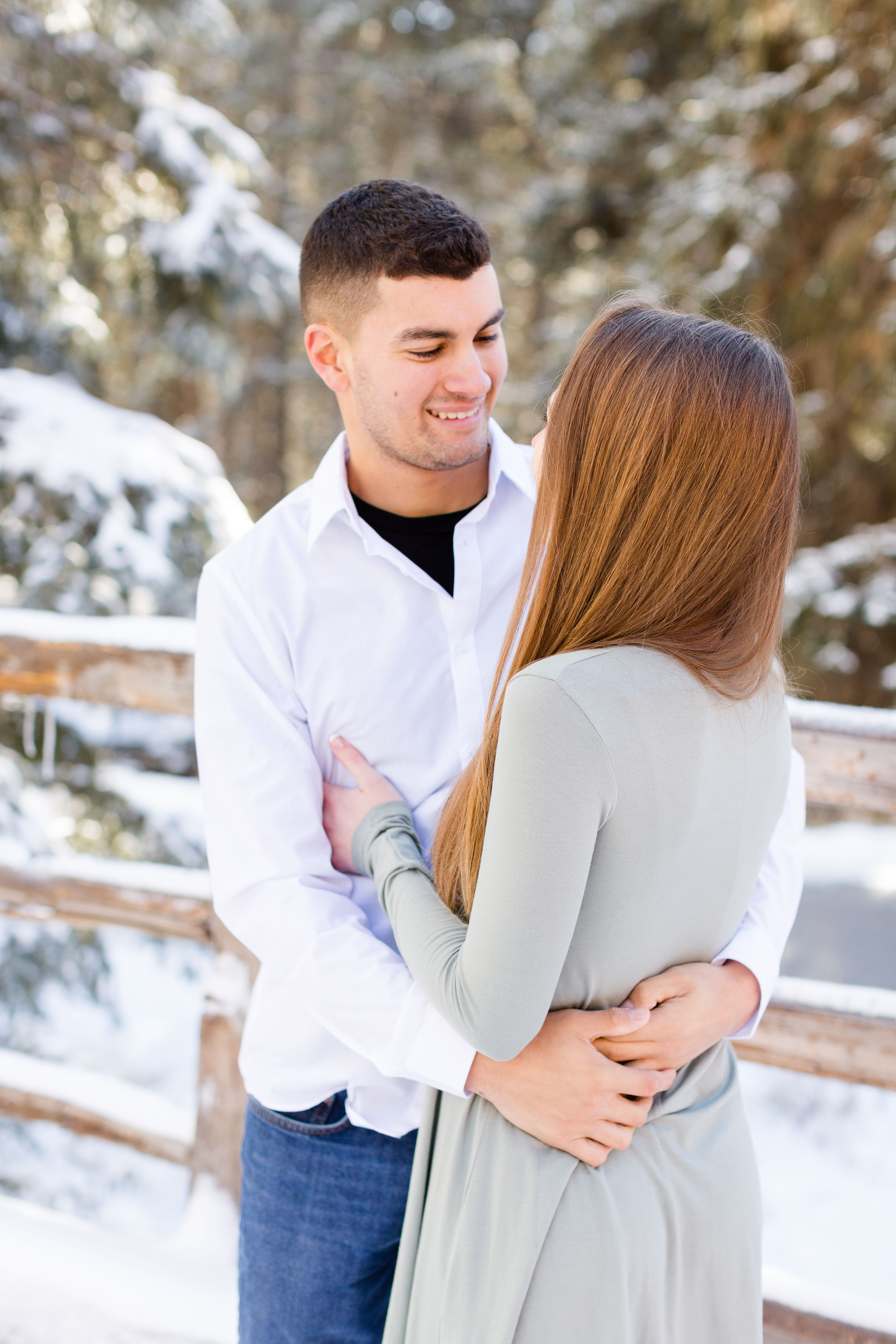 man smiles and hugs his wife in front of snow covered fence and trees