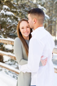 woman smiles as her husband kisses her forehead in front of snow covered fence and pine trees