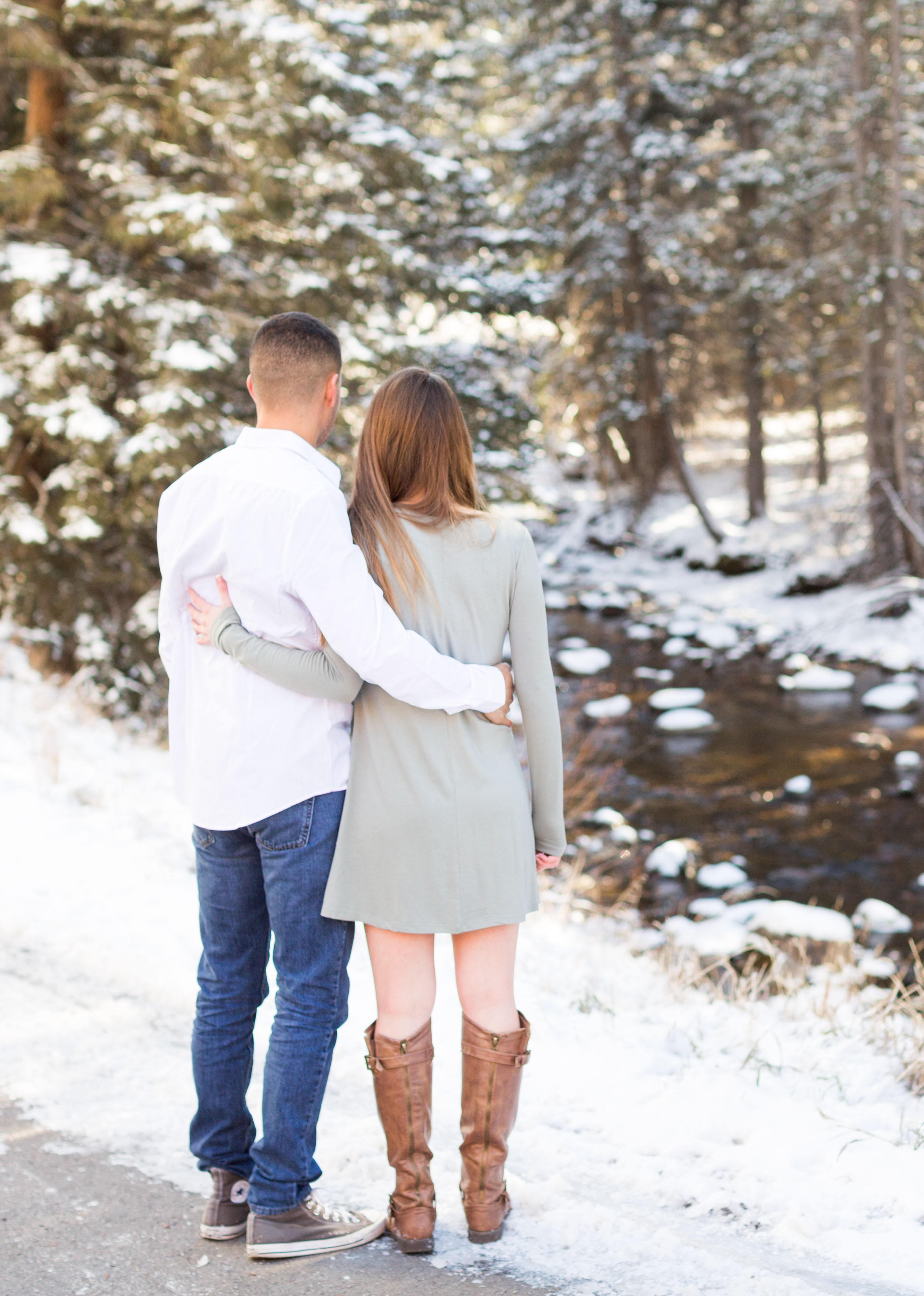 husband and wife hold each other and look at a river on a snowy day in the mountains