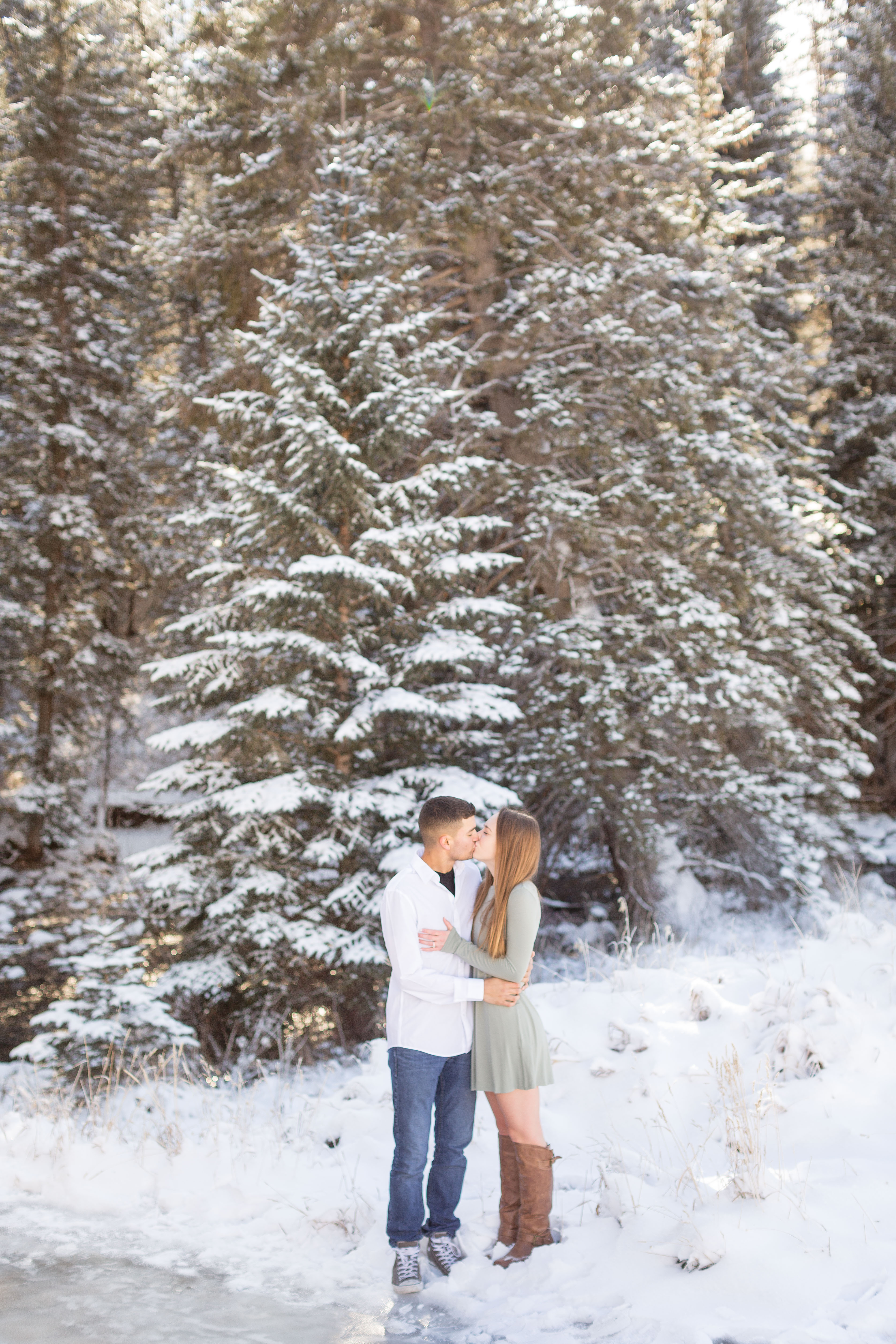couple kisses in front of snow covered pine trees