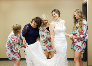 mom and bridesmaids gather around bride and smile as she gets into dress